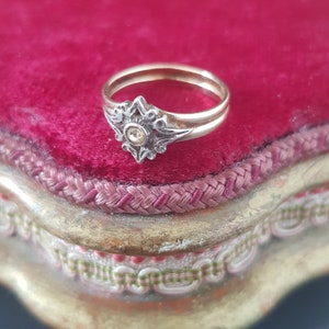 May include: A delicate gold ring with a silver filigree design featuring a small, clear gemstone in the center. The ring is displayed on a red velvet surface.