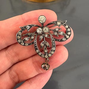 May include: An antique silver brooch with clear gemstones. The brooch has a floral design with a central cluster of stones and a dangling teardrop-shaped stone. The design includes leaf details and a bow-shaped element. The brooch is a vintage piece.