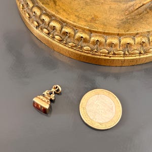 May include: A small, antique gold-colored pendant with a rectangular, reddish-brown stone. The pendant is next to a one Euro coin for size comparison. The background is a dark gray surface with a gold-colored, ornate object.
