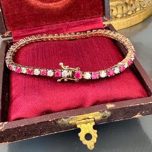 Könnte beinhalten: Ein Tennisarmband mit Rubinen und Diamanten in einer roten Samt-Schmuckschatulle. Das Armband ist aus Gold und hat eine Reihe von runden Rubinen und Diamanten.