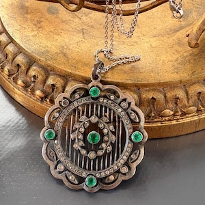 Pode incluir: Colar com pingente prateado com um design circular detalhado. O pingente apresenta um motivo floral central, rodeado por pequenas pedras transparentes e adornado com pedras preciosas verdes. O colar pende de uma corrente delicada.