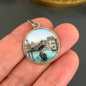 May include: A round silver-toned pendant featuring a hand-painted enamel scene of a Venetian canal with a gondola, a bridge, and buildings. The circular charm has a small metal loop at the top for attachment to a chain or bracelet.