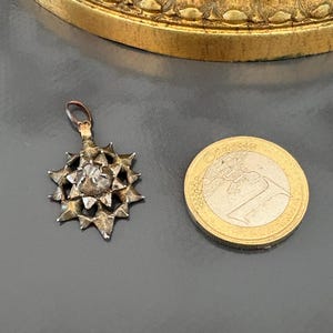 May include: A silver-toned star-shaped pendant featuring a central faceted stone and a small top loop for attachment. The pendant is positioned next to a one-euro coin for scale, which has a diameter of approximately 23.25 millimeters.