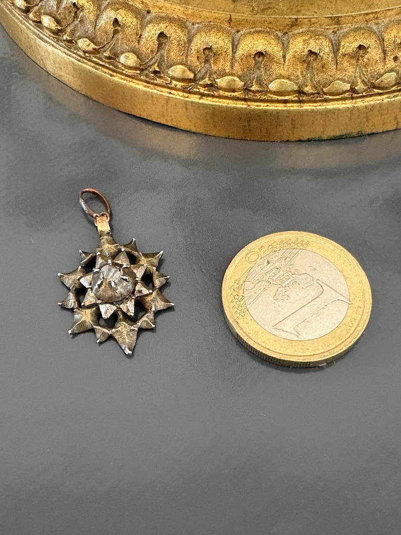 May include: A silver-toned star-shaped pendant featuring a central faceted stone and a small top loop for attachment. The pendant is positioned next to a one-euro coin for scale, which has a diameter of approximately 23.25 millimeters.