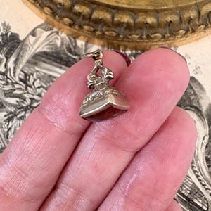 May include: A small, antique silver seal with a decorative handle and a square base. The base appears to be a dark red color. The seal is held in a hand against a backdrop of vintage paper and a gold-toned decorative plate.