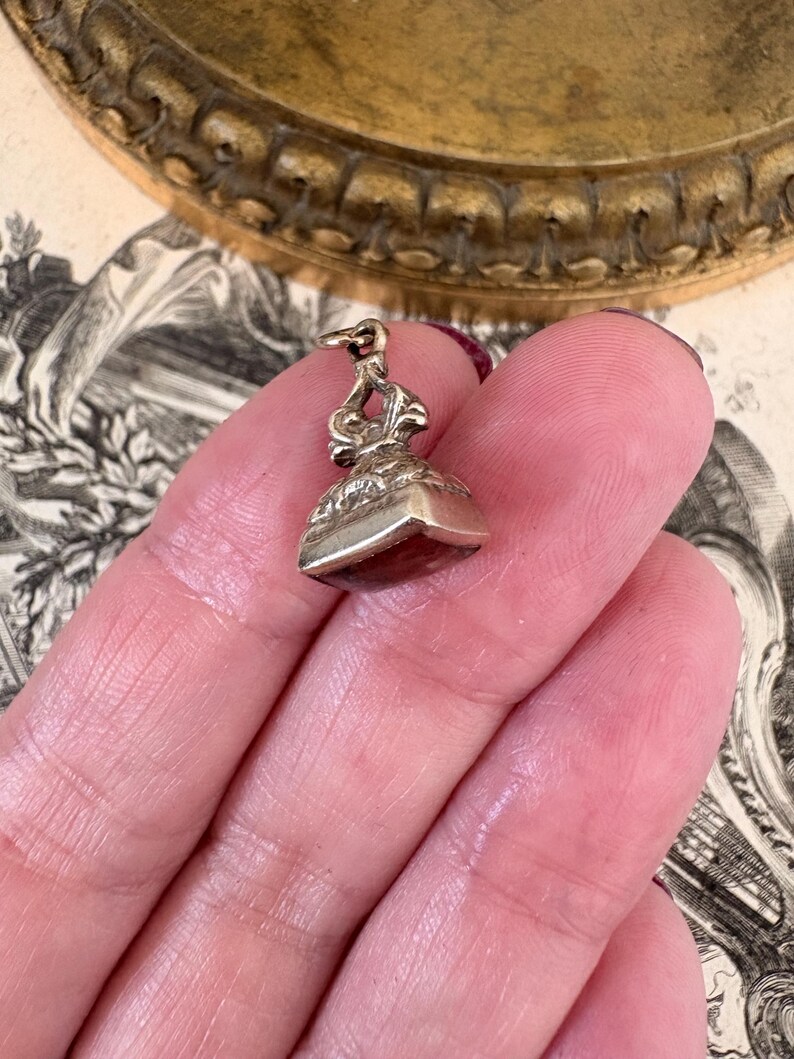 May include: A small, antique silver seal with a decorative handle and a square base. The base appears to be a dark red color. The seal is held in a hand against a backdrop of vintage paper and a gold-toned decorative plate.