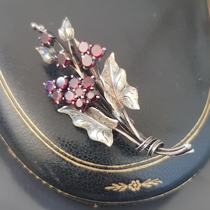 May include: A silver brooch with a bouquet of red garnet flowers and silver leaves. The brooch has a twisted wire stem.