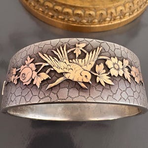 Pode incluir: Pulseira articulada prateada com um design detalhado de pássaros e flores. A pulseira apresenta um pássaro dourado em voo, rodeado por flores e folhas douradas e ouro rosa. O fundo tem um padrão de favo de mel texturizado.