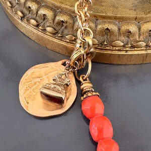May include: A gold-toned charm necklace with a round pendant, a small rectangular charm, and a strand of three red beads. The necklace is hanging in front of a gold-colored, ornate background.
