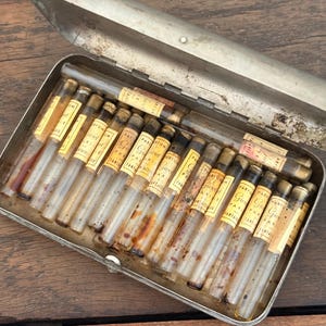 Puede incluir: Un estuche metálico antiguo abierto que contiene numerosos viales de vidrio pequeños. Cada vial tiene una etiqueta amarillenta con texto negro. Los viales están dispuestos ordenadamente dentro del estuche, sugiriendo una colección de perfumes.