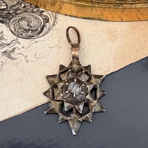 May include: A dark metallic star-shaped pendant featuring a central faceted stone set within a multi-pointed frame. The pendant has a textured, weathered finish and includes a small circular bail at the top for attachment to a chain or cord.