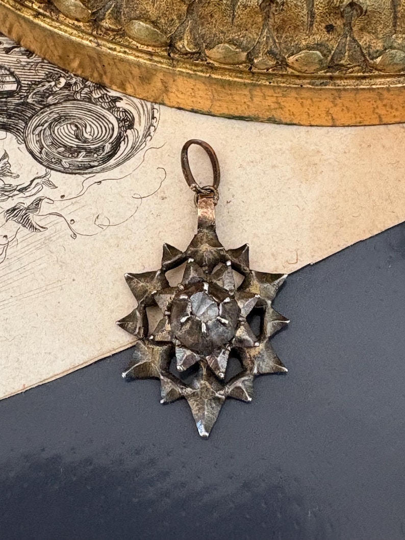May include: A dark metallic star-shaped pendant featuring a central faceted stone set within a multi-pointed frame. The pendant has a textured, weathered finish and includes a small circular bail at the top for attachment to a chain or cord.