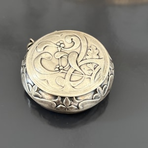 May include: A round, silver-toned pendant with intricate floral and scrollwork designs. The pendant's top surface features a raised, detailed pattern, while the sides are adorned with a repeating floral motif. A small loop is attached for hanging.