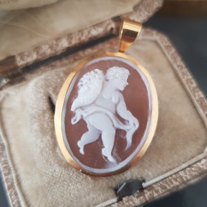 Puede incluir: Un colgante de oro con un camafeo de un querubín con alas, tallado en blanco sobre un fondo marrón. El colgante está en una caja forrada de terciopelo.