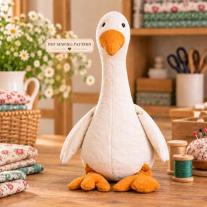 Puede incluir: Un juguete de peluche con forma de ganso de cuerpo blanco, pico naranja y patas palmeadas naranjas. El juguete tiene pequeños ojos negros y una costura central en el pecho. Se ve una etiqueta rectangular con el texto PDF SEWING PATTERN.