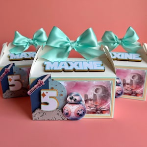 May include: Three favour boxes with a Star Wars theme. The boxes are decorated with a blue and white bow, a space scene, and the name "Maxine".