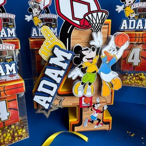 May include: A basketball-themed birthday decoration featuring the number 4 with Mickey Mouse, Donald Duck, and Goofy. The design includes a basketball hoop, a foam finger, and the name "ADAM". Additional decorations include basketball-themed boxes and the text "SLAM DUNK!"