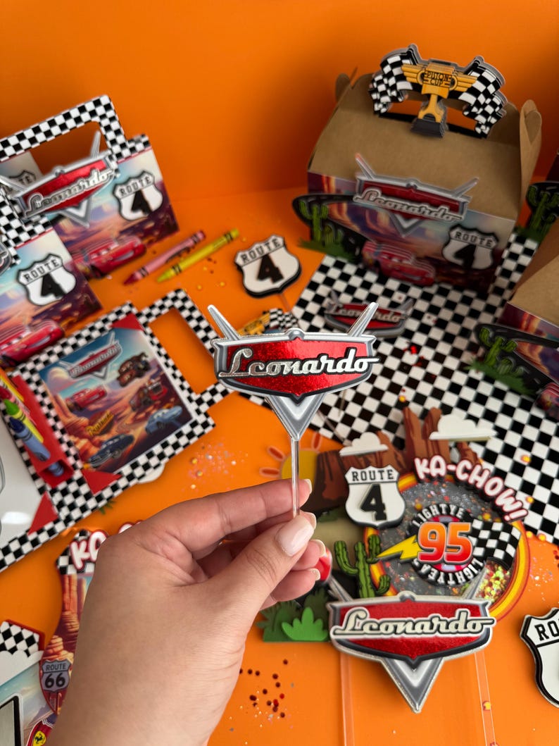 Pode incluir: Um topo de bolo vermelho e prateado com o nome "Leonardo" nele. O topo est&aacute; em um palito e &eacute; cercado por outros topos de bolo com um tema de Carros. Os topos incluem uma placa da Rota 66, um cacto e uma placa de "Ka-Chow!"