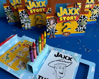 May include: A blue and white coloring book with a picture of Woody from Toy Story on the cover. The book has a handle and a slot for crayons. The text on the cover reads "Jaxx Turns 2!" and there are six crayons in the slot.