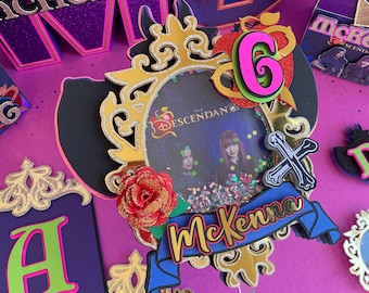 May include: A purple and gold cake topper with a picture frame featuring the Disney Descendants characters. The frame is decorated with a red rose, a blue ribbon, and the name "Mckenna".