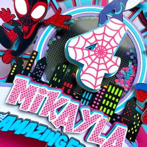 May include: A pink and blue cake topper featuring a Spiderman character, a Spider-Girl character, and the number 4. The cake topper also includes a cityscape and the name "Mikayla" with the words "Amazing Friend" underneath.