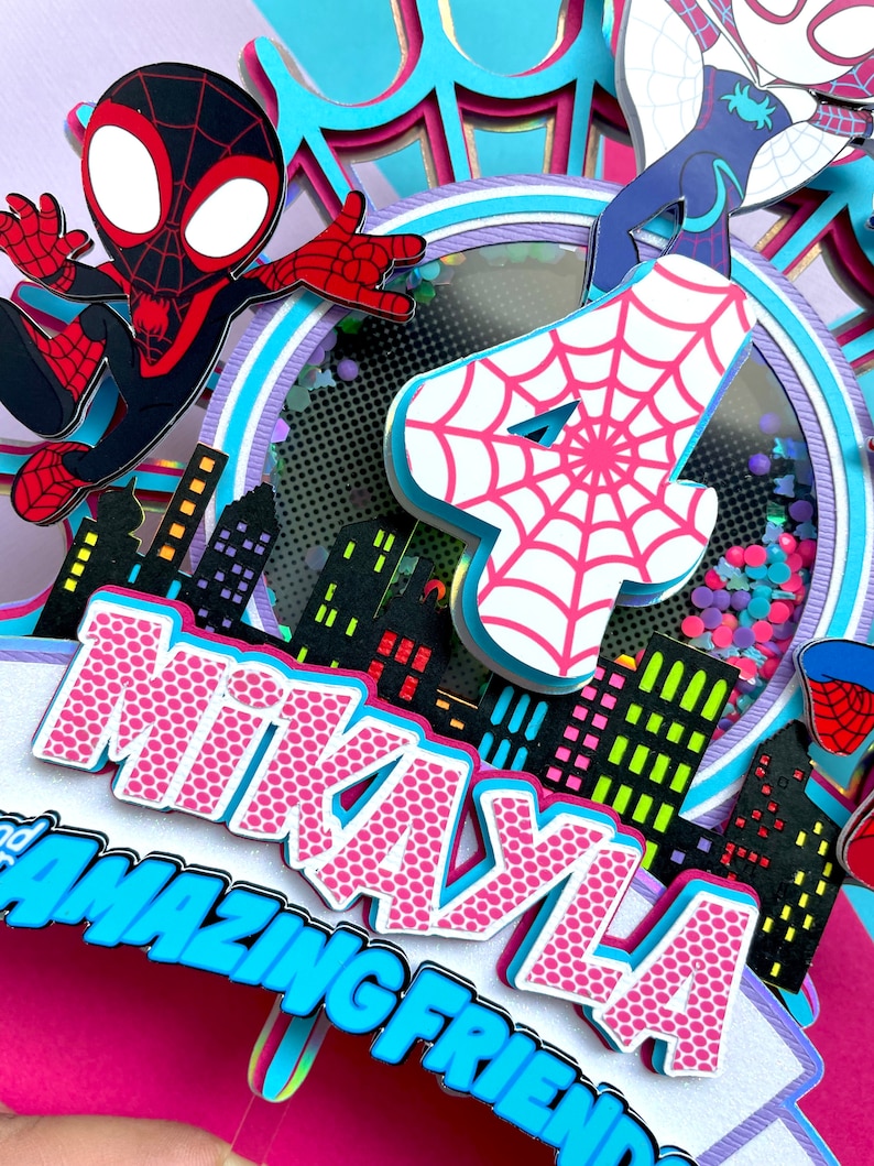 May include: A pink and blue cake topper featuring a Spiderman character, a Spider-Girl character, and the number 4. The cake topper also includes a cityscape and the name "Mikayla" with the words "Amazing Friend" underneath.