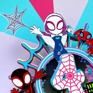May include: A colourful cake topper featuring Spider-Man and Spider-Gwen characters. The topper includes a pink number 4 with a white spiderweb design, a red and black Spider-Man figure, a white Spider-Gwen figure, and a red spider. The topper is ideal for a Spider-Man themed birthday party.