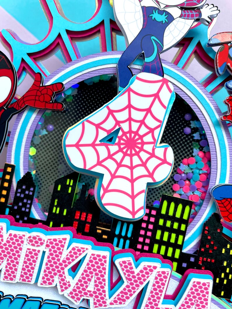May include: A pink and white cake topper with a spiderweb design. The topper features a pink number four, a spiderweb design, and a cityscape. The cake topper also includes the name "KAYLA" in pink letters.