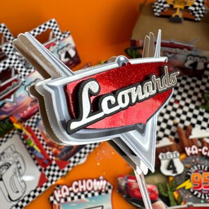 Pode incluir: Um topo de bolo vermelho e prateado com o nome "Leonardo" em letras pretas. O topo tem a forma de um carro e tem um padr&atilde;o xadrez na parte inferior. Est&aacute; em um palito de pl&aacute;stico transparente.