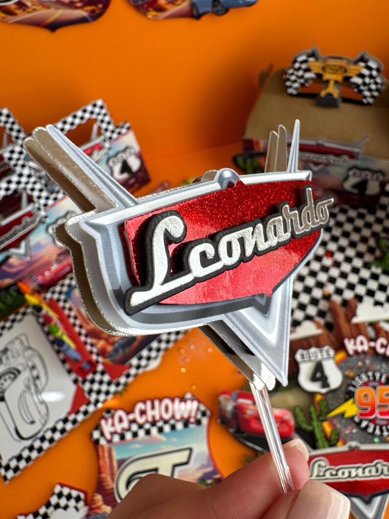 Pode incluir: Um topo de bolo vermelho e prateado com o nome "Leonardo" em letras pretas. O topo tem a forma de um carro e tem um padr&atilde;o xadrez na parte inferior. Est&aacute; em um palito de pl&aacute;stico transparente.