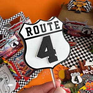 Pode incluir: Topper de bolo branco e preto com uma placa branca que diz "ROUTE 4" em texto preto. A placa tem a forma de uma placa de estrada cl&aacute;ssica.