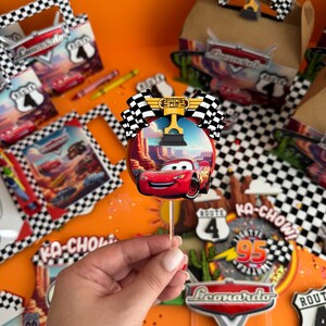 Pode incluir: Um topo de bolo de carro de corrida vermelho com uma bandeira quadriculada e um trof&eacute;u. O topo de bolo est&aacute; em um palito e &eacute; cercado por outras decora&ccedil;&otilde;es de festa, incluindo uma placa que diz "Ka-Chow!" e uma placa que diz "Rota 95".