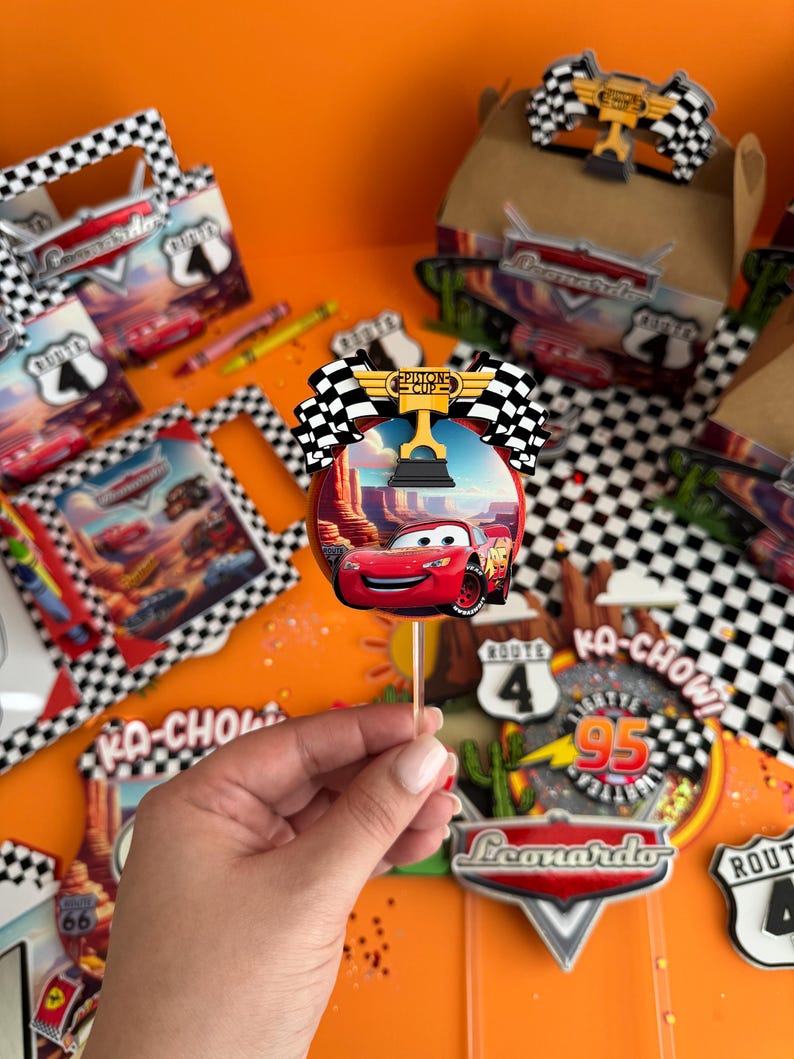Pode incluir: Um topo de bolo de carro de corrida vermelho com uma bandeira quadriculada e um trof&eacute;u. O topo de bolo est&aacute; em um palito e &eacute; cercado por outras decora&ccedil;&otilde;es de festa, incluindo uma placa que diz "Ka-Chow!" e uma placa que diz "Rota 95".