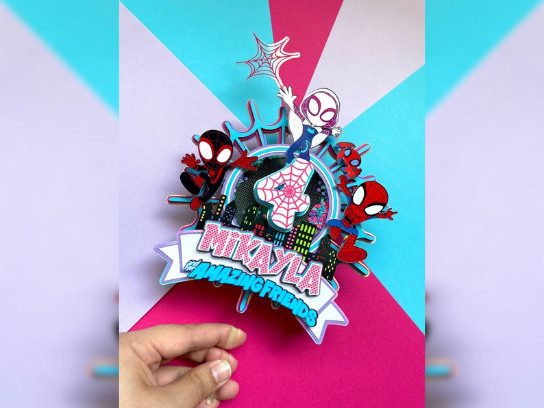May include: A pink and blue cake topper with the name "Mikayla" and the words "and the Amazing Friends" in a white banner. The topper features a number 4 with a spiderweb design and four cartoon Spider-Man characters.