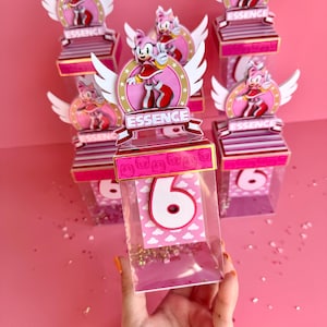 May include: Pink and white party favor boxes with a cartoon character on the top. The boxes are decorated with gold glitter and the word "ESSENCE". The boxes are filled with gold glitter and a white number six.