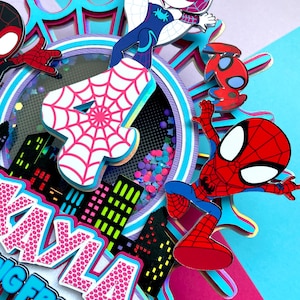 May include: A pink and blue multi-layered cake topper featuring Spider-Man, Spider-Gwen, and a number 4. The cake topper is decorated with a cityscape, glitter, and a spiderweb design. The text "Kayla's Big Friend" is visible on the bottom of the topper.