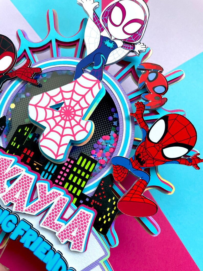 May include: A pink and blue multi-layered cake topper featuring Spider-Man, Spider-Gwen, and a number 4. The cake topper is decorated with a cityscape, glitter, and a spiderweb design. The text "Kayla's Big Friend" is visible on the bottom of the topper.