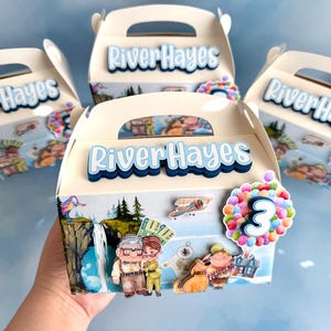 Könnte beinhalten: Drei Party-Geschenkboxen im Up-Thema mit blau-weißem Design. Die Boxen zeigen den Namen "River Hayes" und ein Cartoon-Bild eines Paares mit einem Heißluftballon. Die Boxen sind mit einem blau-weißen Design verziert und haben einen Griff oben.