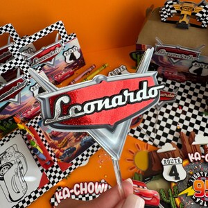 Pode incluir: Um topo de bolo vermelho e prateado com o nome "Leonardo" em letras prateadas com glitter. O topo tem a forma de um raio e um fundo vermelho. O topo est&aacute; em um palito de pl&aacute;stico transparente.