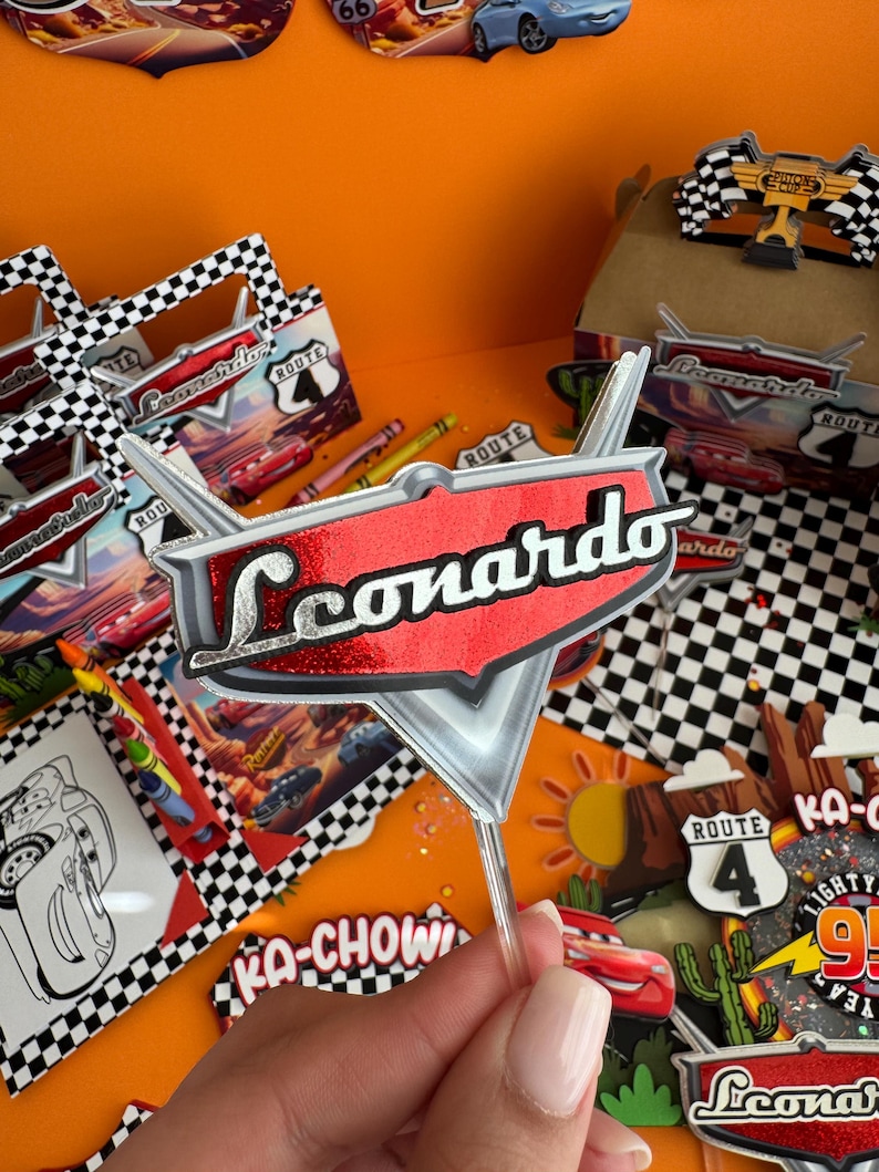 Pode incluir: Um topo de bolo vermelho e prateado com o nome "Leonardo" em letras prateadas com glitter. O topo tem a forma de um raio e um fundo vermelho. O topo est&aacute; em um palito de pl&aacute;stico transparente.