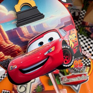 Pode incluir: Topper de bolo de carro de corrida vermelho de desenho animado com um rosto sorridente. O carro tem rodas pretas com o texto "YEAR" e "LIGHTYEAR" nos pneus. O carro est&aacute; em um palito e est&aacute; pronto para ser colocado em um bolo.