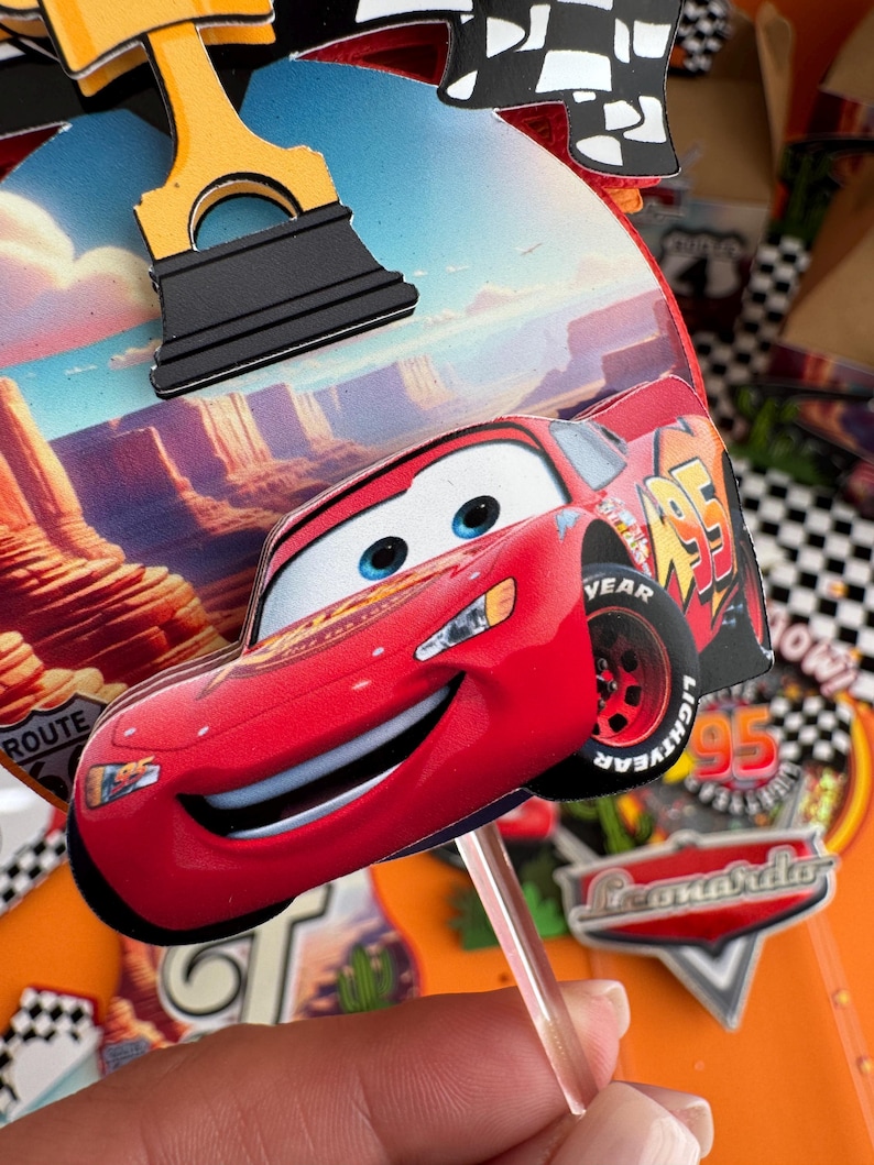 Pode incluir: Topper de bolo de carro de corrida vermelho de desenho animado com um rosto sorridente. O carro tem rodas pretas com o texto "YEAR" e "LIGHTYEAR" nos pneus. O carro est&aacute; em um palito e est&aacute; pronto para ser colocado em um bolo.