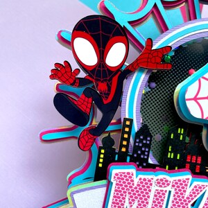 May include: A 3D papercraft featuring a cartoon Spider-Man character in a black and red suit, with a cityscape backdrop and the name "Mike" in pink letters with a white polka dot pattern.