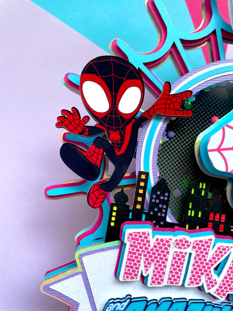 May include: A 3D papercraft featuring a cartoon Spider-Man character in a black and red suit, with a cityscape backdrop and the name "Mike" in pink letters with a white polka dot pattern.