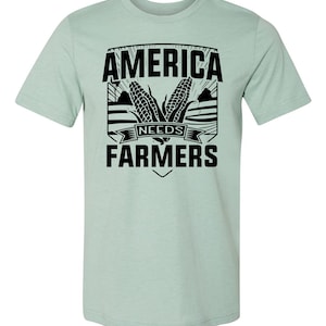 May include: A light blue t-shirt with a black graphic design that says "America Needs Farmers". The graphic features two ears of corn and a stylized field.