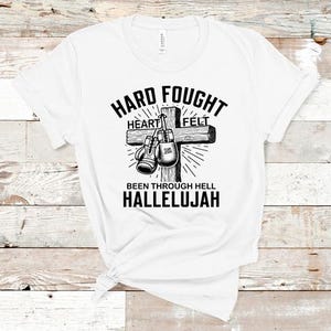 May include: White t-shirt with a black graphic design of a cross with boxing gloves hanging from it. The text reads "Hard Fought Heart Felt Been Through Hell Hallelujah".