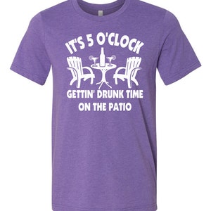 May include: Purple t-shirt with white text that reads "It's 5 o'clock Gettin' Drunk Time On The Patio". The text is accompanied by a graphic of two chairs, a table, and drinks.
