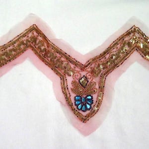 May include: A pink, V-shaped fabric applique with intricate gold and bronze beadwork and sequins. The center features a cluster of blue and clear gemstones. The applique is likely for embellishing clothing or accessories.