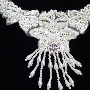 May include: White lace applique with pearl embellishments and a beaded fringe. The applique features a floral design with a central butterfly motif.