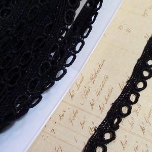 May include: Close-up of black lace trim with a scalloped edge. The lace is made of a delicate, open-weave material. The trim is displayed on a vintage paper background with handwritten text.